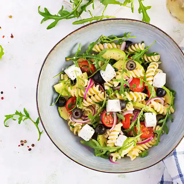 A large glass or white ceramic bowl filled with colorful bow tie pasta salad, mixed with cherry tomatoes, cucumber, carrots, bell peppers, spinach, and red onion, lightly dressed and garnished with fresh basil and black pepper, served on a wooden table in natural light.