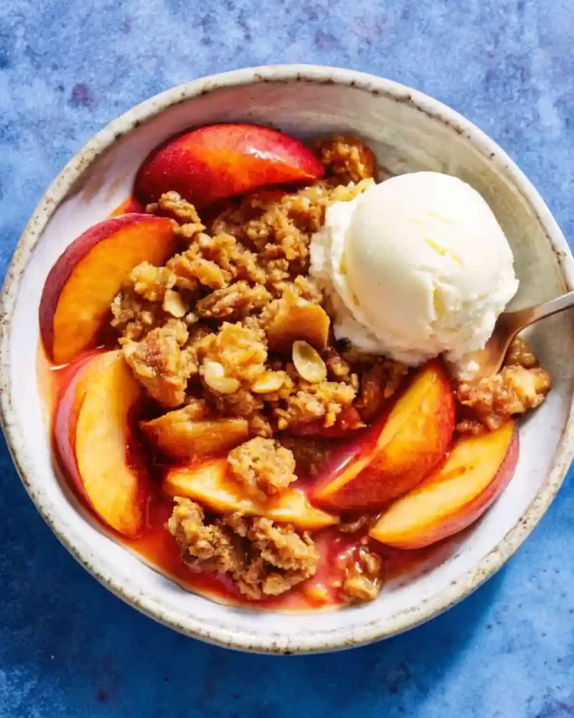 Peach Crisp Recipe You’ll Love – Easy & Crispy Dessert | QuikyKitchen Warm peach crisp served in a ceramic bowl with vanilla ice cream on top, blue background