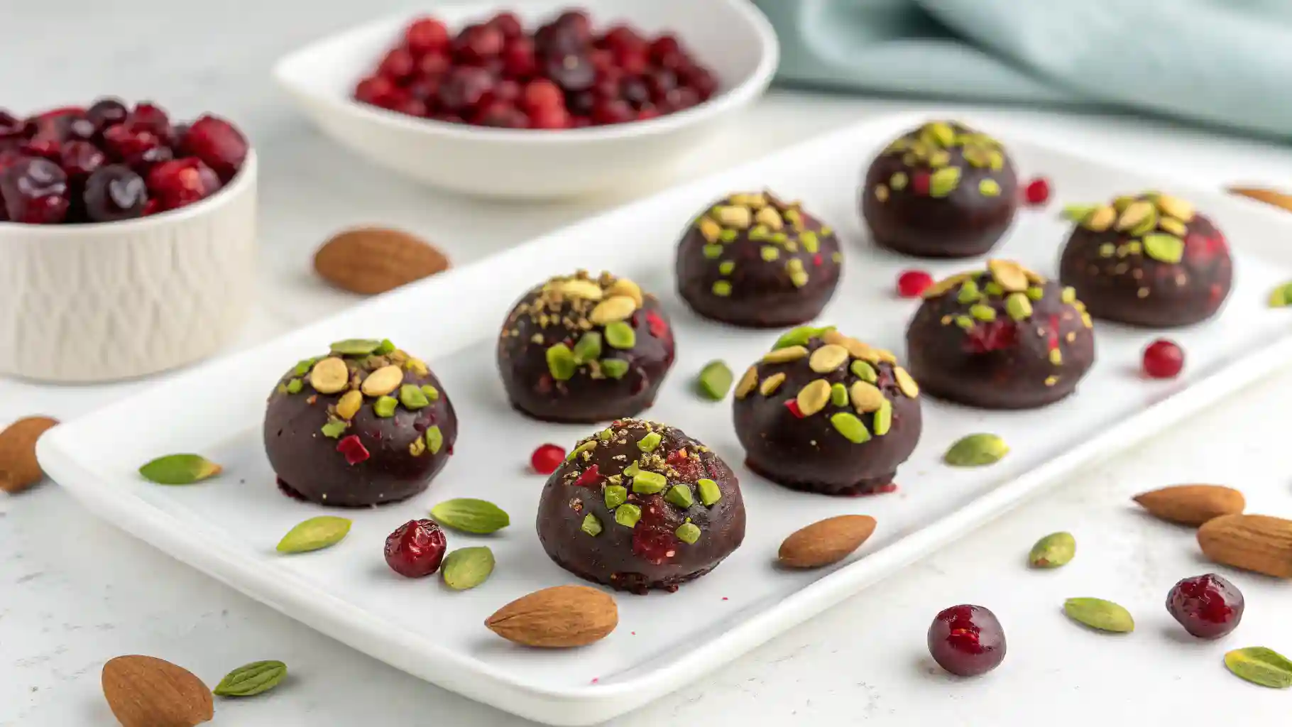 Dark chocolate truffles topped with crushed pistachios and almonds, served on a white tray surrounded by cranberries and nuts