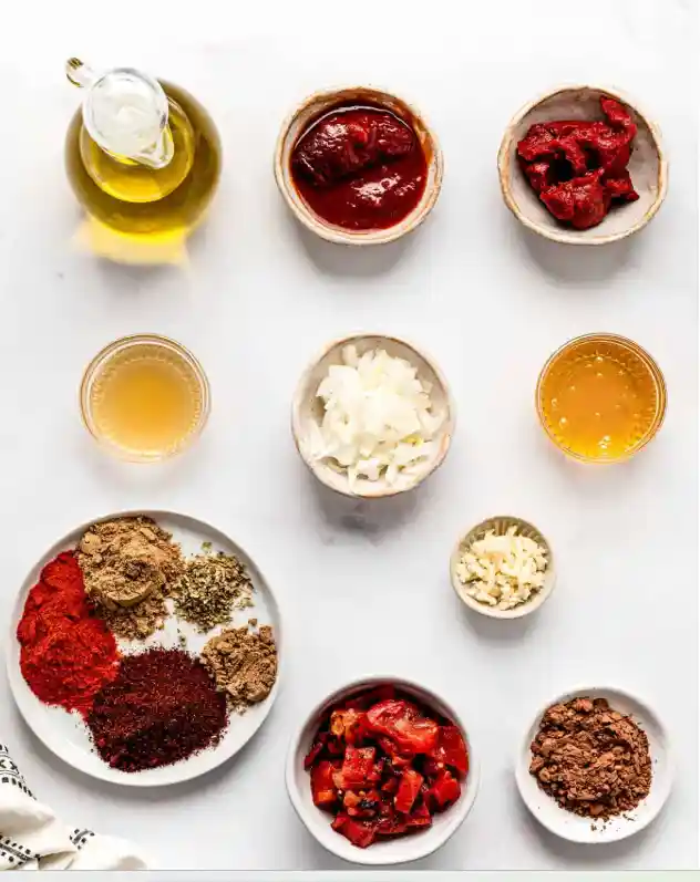 Hello Fresh Tex Mex Paste Recipe (Easy Copycat) Overhead view of ingredients for homemade Tex Mex paste, including olive oil, tomato paste, chipotle in adobo, chopped onion, garlic, roasted peppers, broth, honey, and a mix of chili powder, paprika, cumin and spices on a white background