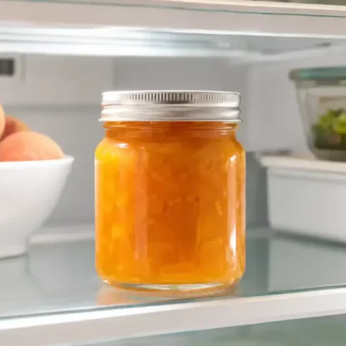Easy Peach Jam Recipe Without Pectin Jar of homemade peach jam recipe stored on a refrigerator shelf beside fresh peaches